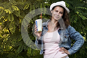 Portrait of cute attractive girl in hat with glass of cold drink with straw in park nature