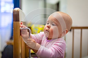 Portrait of cute baby girl with blue eyes is standing in crib. Adorable infant is standing up in cot and is interest