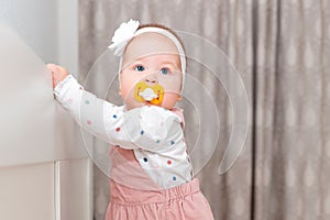 Portrait of cute baby girl in a dress, with a pacifier in her mouth and with a headband on her head, stands near the crib. The