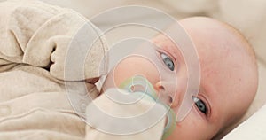 Portrait of a cute baby with a pacifier in his mouth, lying on a white blanket, in the white clothes, sobbing.