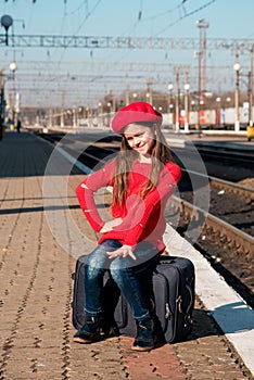 Portrait of a cute beautiful joyful girl sitting on a suitcase and waiting for a train at the train station