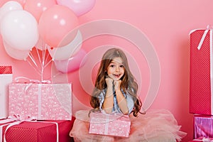 Portrait cute birthday kid expressing to camera suround big giftboxes, balloons  on pink background. Amazing