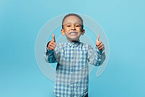 Portrait of cute black boy showing thumbs up and smiling at camera while posing over blue background, studio shot