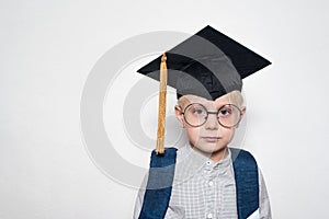 Portrait of a cute blond boy in big glasses, academic hat and a school bag. White background. Place for text