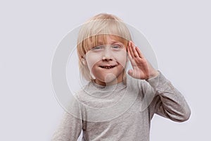 Portrait of cute blond boy on white background. Child holds hand of face and grins