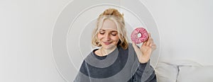 Portrait of cute blond girl holding pink doughnut with sprinkles on top, showing her favourite food