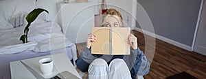 Portrait of cute blond woman, sits in front of digital camera and laptop in her room, covers face with notebook journal