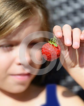 Portrait of cute blonde Caucasian girl is holding a red berry in her hand. Girl is smiling. The berry is closed up. Girls is blurr