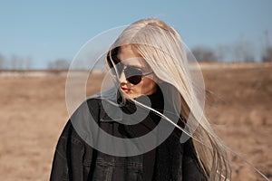 Portrait of cute blonde girl in the field, wearing sunglasses and black jacket.