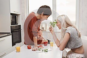 Portrait of cute blonde girl holding salary and talking to her girlfriend while she sits on kitchen table and laughs