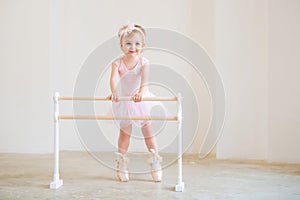 Portrait of a cute blue-eyed baby ballerina in a pink leotard and big pointe shoes standing near the ballet barre