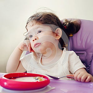 Portrait of cute bored Caucasian child kid girl sitting in high chair eating cereal with spoon early morning