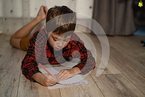 Portrait of cute boy laying on the floor at home writing christmas letter to santa with red pencil. Image with selective focus