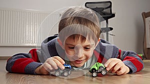 Portrait of cute boy lying on floor and playing with two toy cars. Children playing alone, development and education.