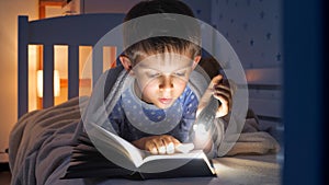 Portrait of cute boy in pajamas holding flashlight and reading interesting book. Children education, development, secrecy, privacy