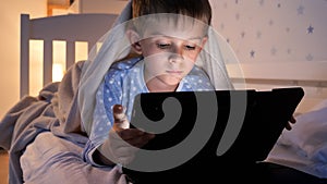 Portrait of cute boy in pajamas lying in bed and browsing internet on tablet computer. Children education, development, kids using