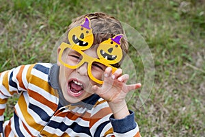 Portrait of cute boy wearing funny carnival orange glasses with pumpkins and grimacing in park. Happy Halloween.