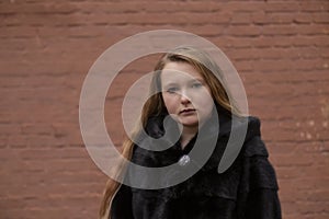 Portrait of cute brunette teen girl in black fur coat on the background of brown brick wall . Cosiness, fashion, style