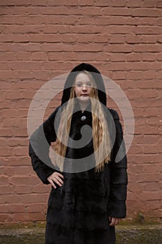 Portrait of cute brunette teen girl in black fur coat on the background of brown brick wall . Cosiness, fashion, style