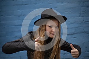 Portrait of cute brunette teen girl in Leather Jacket and black hat showing big thumbs up, on the background of blue brick wall .