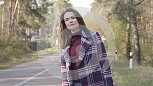 Portrait of cute Caucasian brunette girl hitchhiking on small road in the forest. Positive young woman in plaid jacket