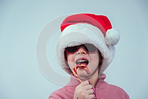 Portrait of a cute Caucasian girl in a Santa Claus hat and sunglasses eating a lollipop on a white background.