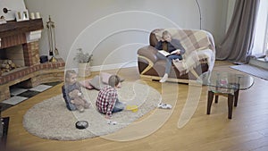 Portrait of cute Caucasian girl sitting on couch with book as her younger sister and brother playing on the floor. Elder