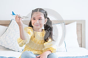 Portrait of cute Caucasian little girl with pony tails hair sitting on bed at home and playing with airplane model toy, holding