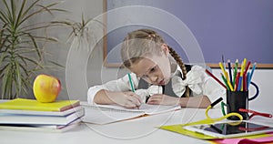 Portrait of cute Caucasian schoolgirl with pigtails sitting at the table and writing in exercise book. Diligent