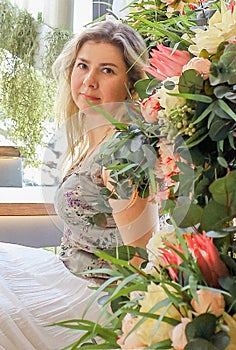 Portrait of a cute Caucasian woman with blond hair, top and white maxi skirt sitting near flowers.