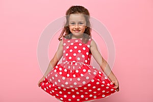 Portrait of cute charming girl in a pink dress with white polka dots posing onpink background cute smiling and holding the bottom
