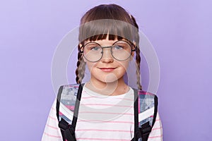 Portrait of cute charming schoolgirl in glasses wearing backpack standing looking at camera with smile, with calm facial