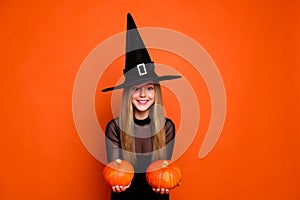 Portrait of cute cheerful wizard girl hands hold festive pumpkins empty space isolated on orange color background