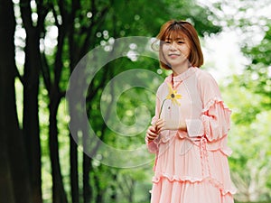 Portrait of a cute Chinese girl in pink dress holding wild flower and smiling at camera in summer forest. Outdoor fashion portrait