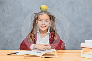 Portrait of cute clever girl with apple on her head. Sitting from a stack of books at the table, a copy of the space