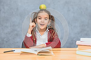 Portrait of cute clever girl with apple on her head. Sitting from a stack of books at the table, a copy of the space