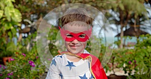 Portrait of a cute funny child boy in a superhero costume, in a red cloak and red mask, plays a superhero, demonstrates