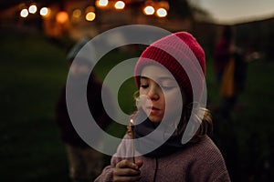 Portrait of cute girl blowing out flame on a burning stick. Family having a barbecue in the garden on an autumn evening.