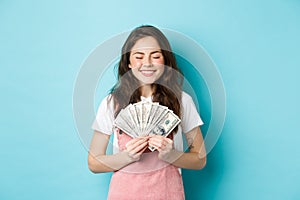 Portrait of cute girl smiling with satisfaction, holding money and looking pleased, winning prize in dollar bills