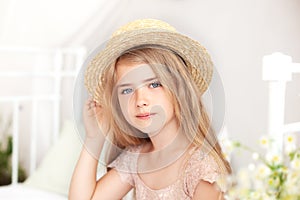 Portrait of a cute girl in a straw hat. Thoughtful little blonde with long hair sits on the bed in a bright children`s room. Child