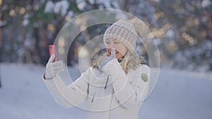 Portrait of cute girl in warm winter clothes talking for video chat using mobile phone and rejoice. Young woman posing