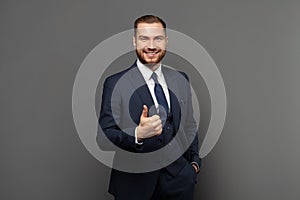 Portrait of cute guy successful businessman wearing suit showing thumb up against grey studio wall background.