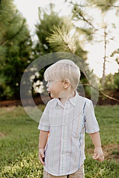 Portrait of Cute Handsome Blond Little Kid Boy in Colorful Striped Button Down Shirt Walking Outside in the Grass Watching the Sun