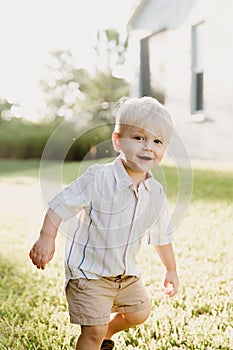 Portrait of Cute Handsome Blond Little Kid Boy in Colorful Striped Button Down Shirt Walking Outside in the Grass Watching the Sun