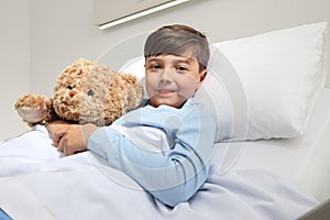 Portrait of cute happy child alone in hospital room lying in bed with teddy bear looking at camera and smiling with copy space