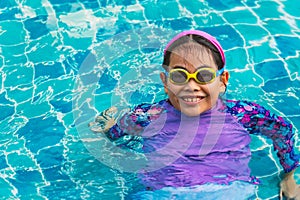Portrait of a cute and healthy Asian child girl with goggles swimming in a swimming pool, smiling, eyes looking at the camera,