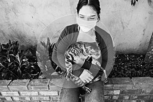 Portrait of a cute house cat lying with her owner at home. A young girl in a medical mask petting a kitten. Black and white