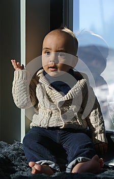 Portrait of a cute infant in beige sweater sitting on window sill