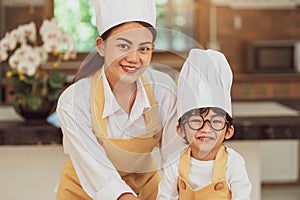 Portrait cute little Asian boy chef with eyeglasses and his mother looking to camera in home cooking kitchen happily and funny.