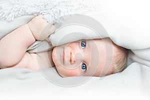 Portrait of cute little baby with blue eyes laying under white blanket and smiling